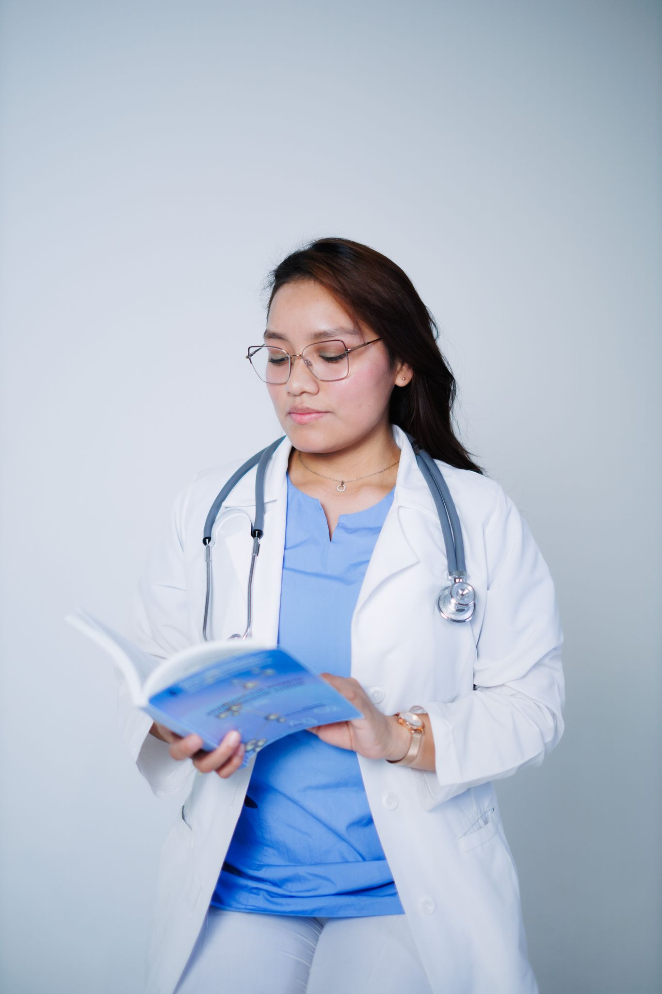 A doctor in a white coat and glasses reads a medical book against a plain background.