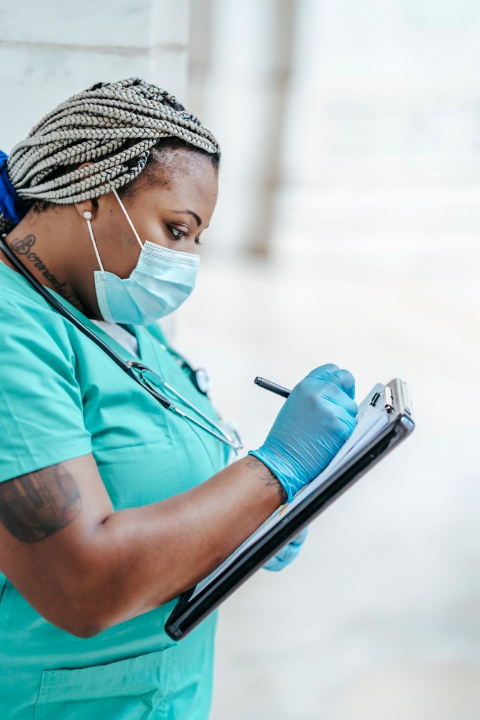 Side view of tattooed mature ethnic female medic in uniform taking notes on paper on clipboard